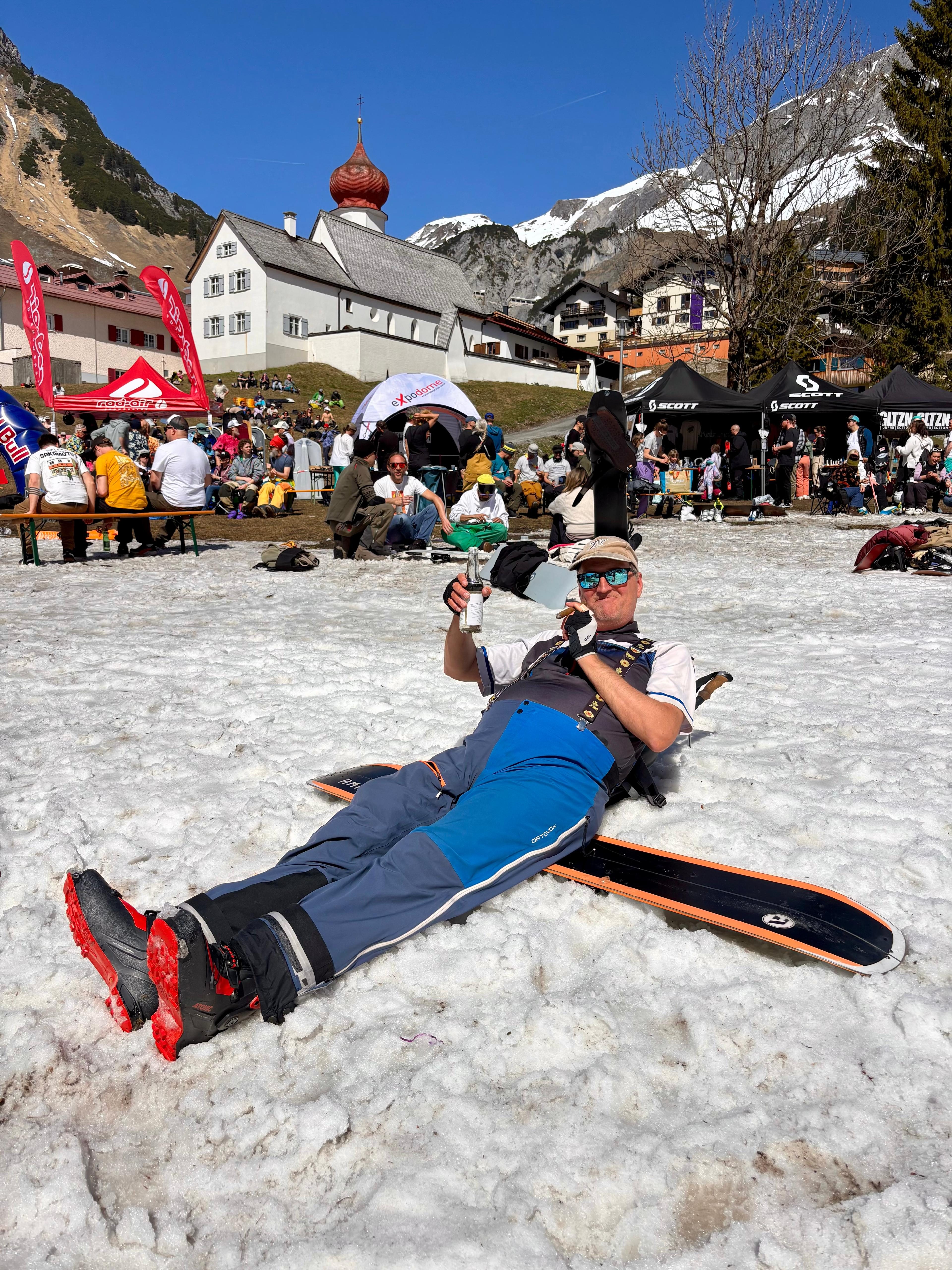 Mann liegt entspannt mit Snowboard im Firn, Stuben Kirche und Dorf im Hintergrund