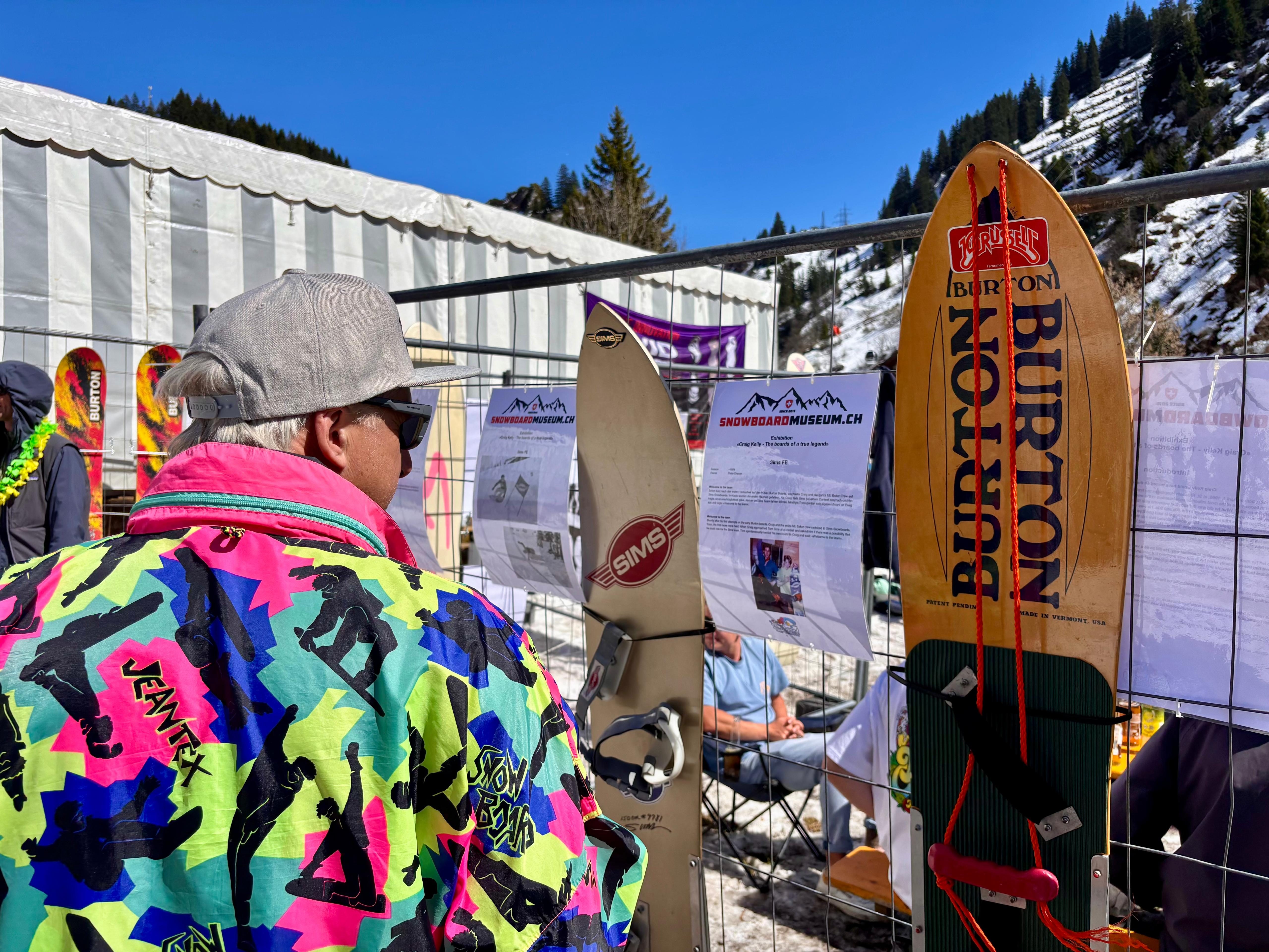 Mann in Neonjacke betrachtet Vintage-Snowboards am Zaun – Snowboard Museum mit Burton und Sims Boards