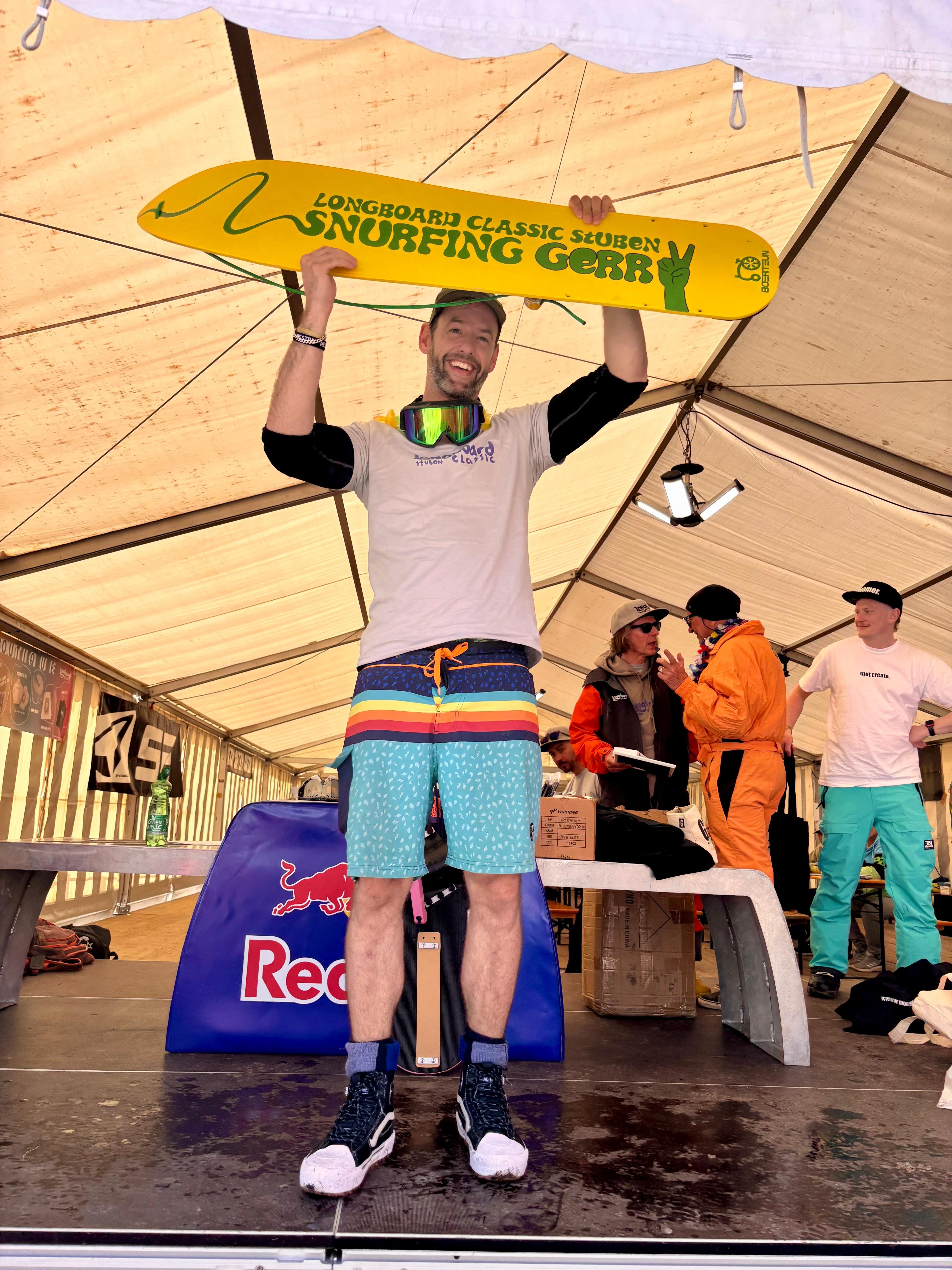 Mann in Shorts hält gelbes Longboard Classic Stuben Snurfing Gear Board über den Kopf