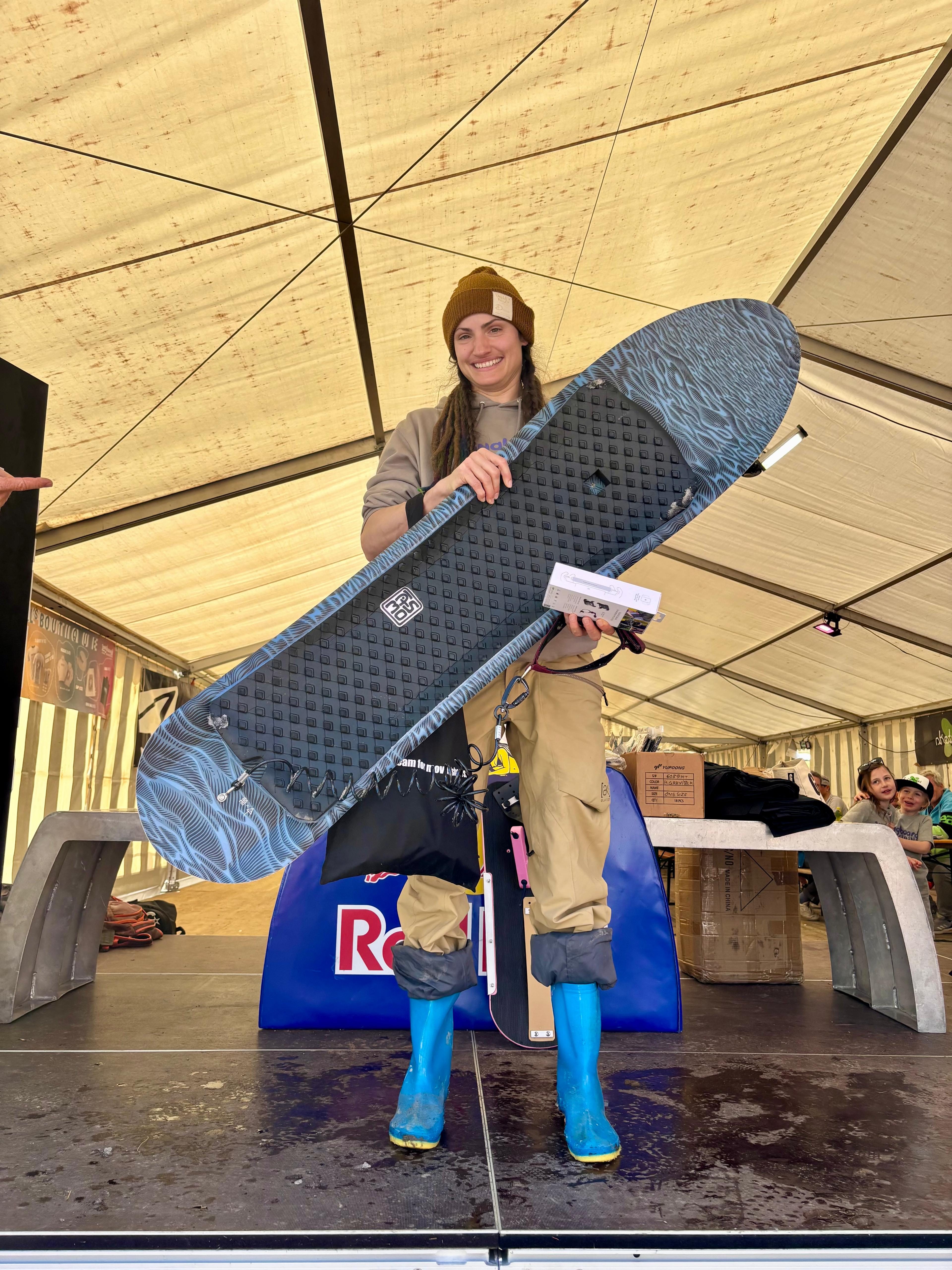 Frau auf der Bühne mit dunklem Splitboard und Urkunde, blaue Gummistiefel